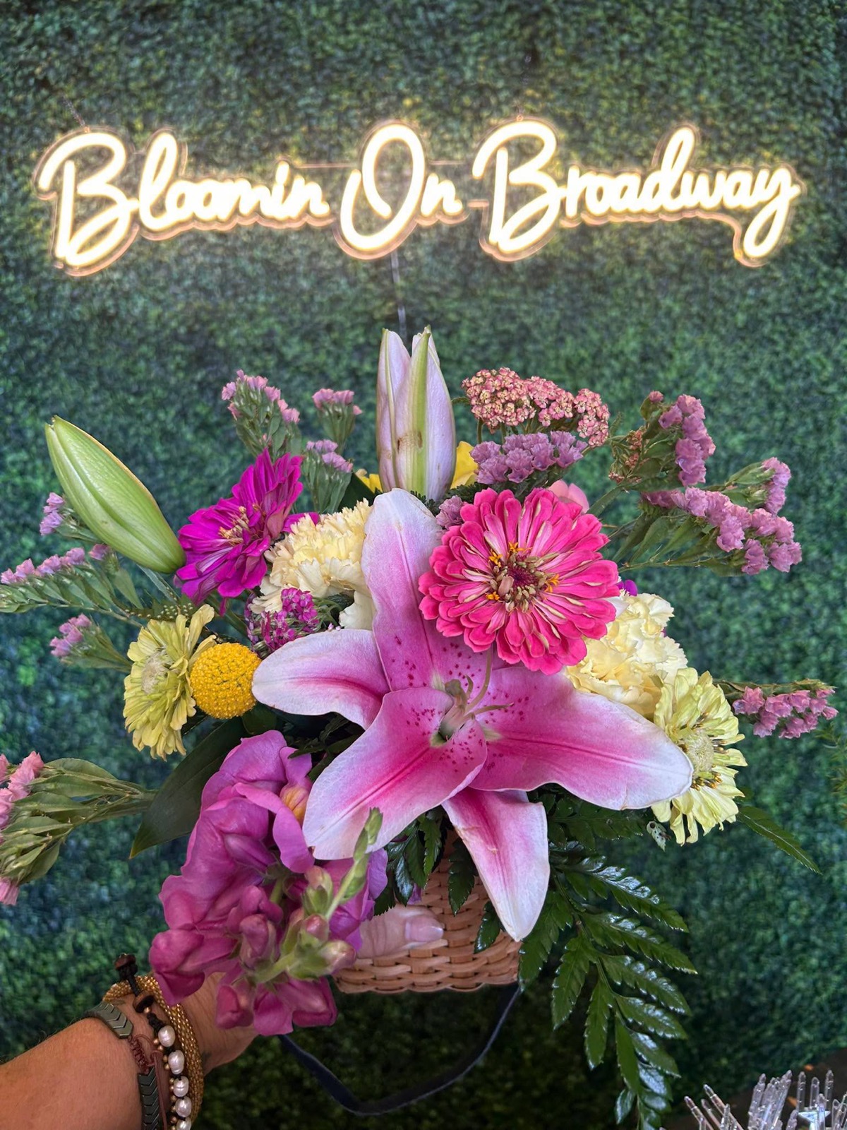 A basket arrangement of pink lilies, zinnias and carnations beneath the Bloomin On Broadway neon sign